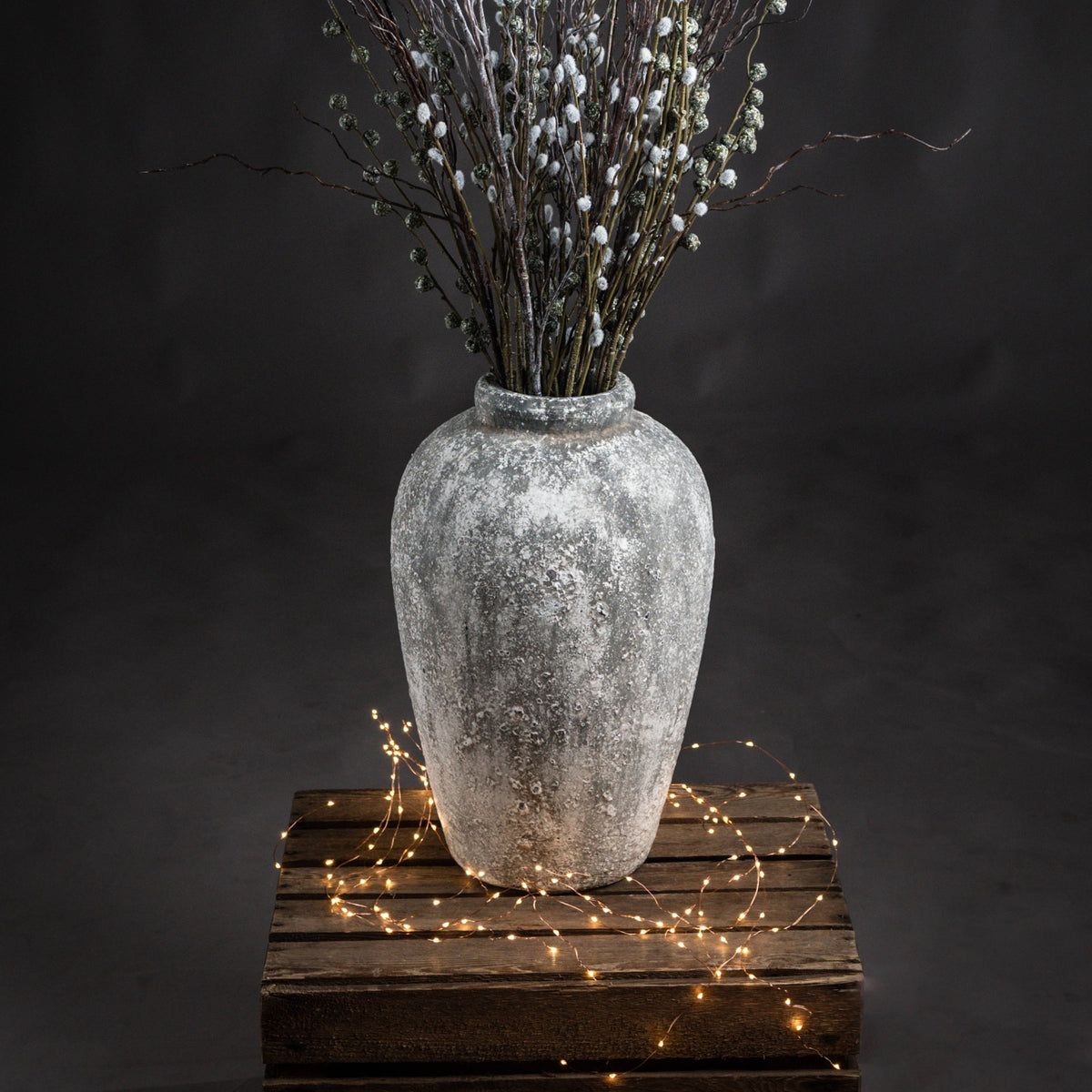 Patchneck Vase filled with tall decorative branches on a wooden crate, with string lights around the base against a dark backdrop.