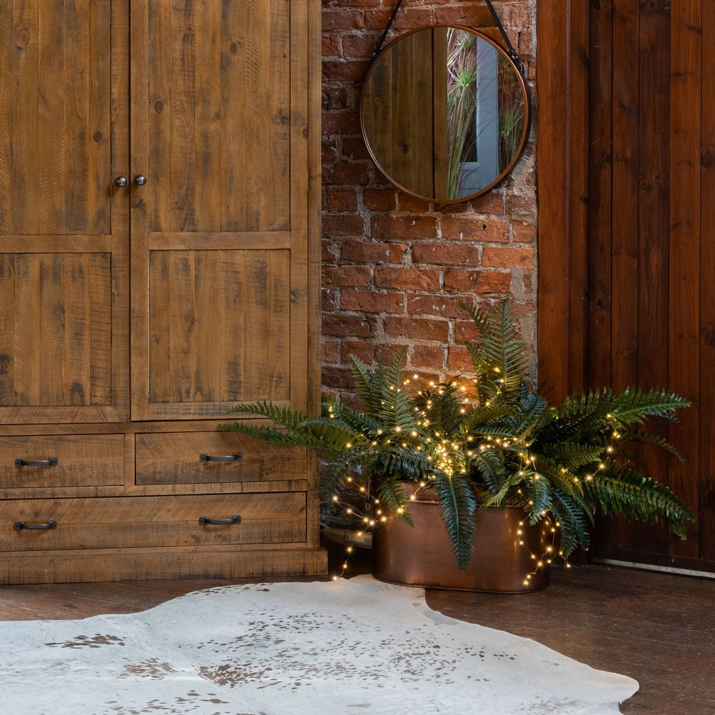 Lit string lights arranged through fern foliage in a copper-toned planter beside a wooden cabinet, beneath a round mirror on a brick wall.