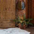 Lit string lights arranged through fern foliage in a copper-toned planter beside a wooden cabinet, beneath a round mirror on a brick wall.