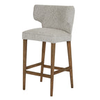 Three-quarter front view of the mist-grey bar stool, showing its winged back, piped seat and tapered warm-brown legs with a footrest.