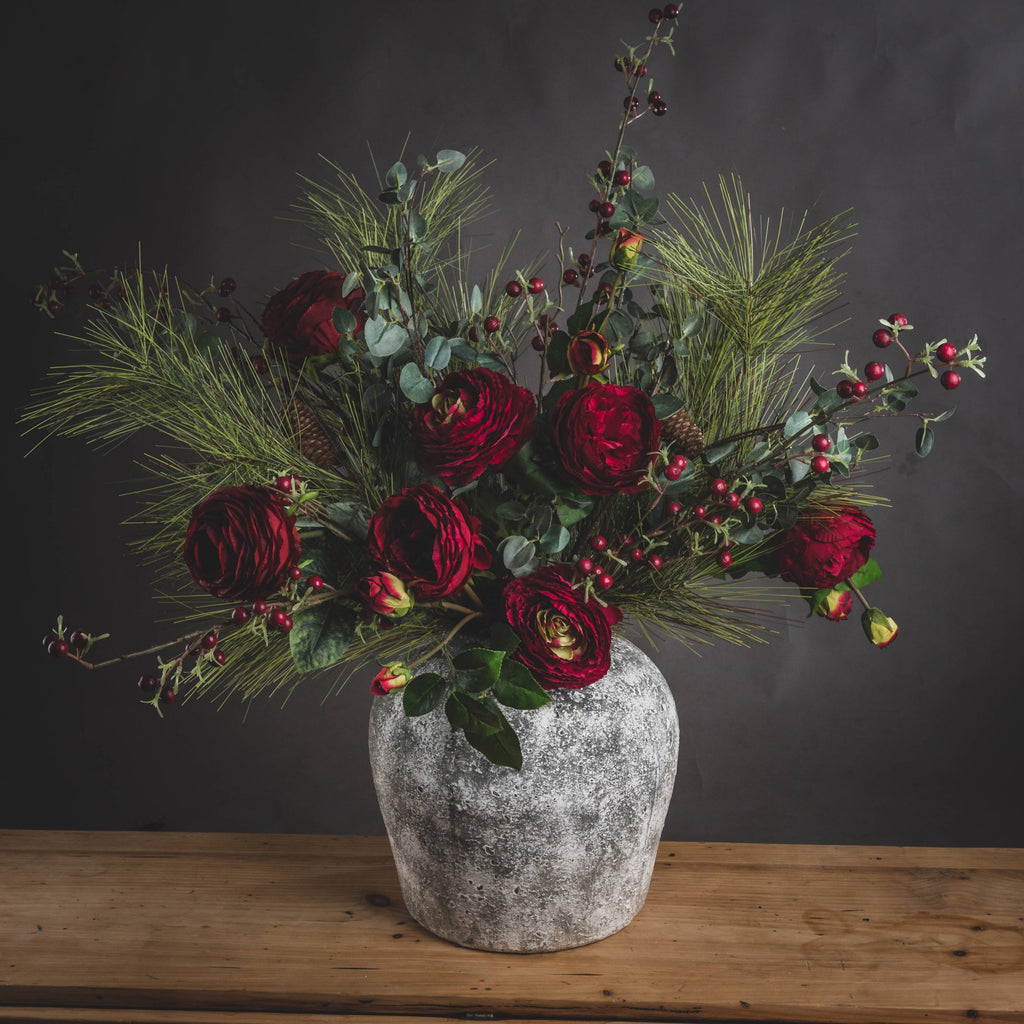 Pitted Round Vase on a wooden table with a spreading arrangement of deep red flowers, berries and green foliage against a dark wall.
