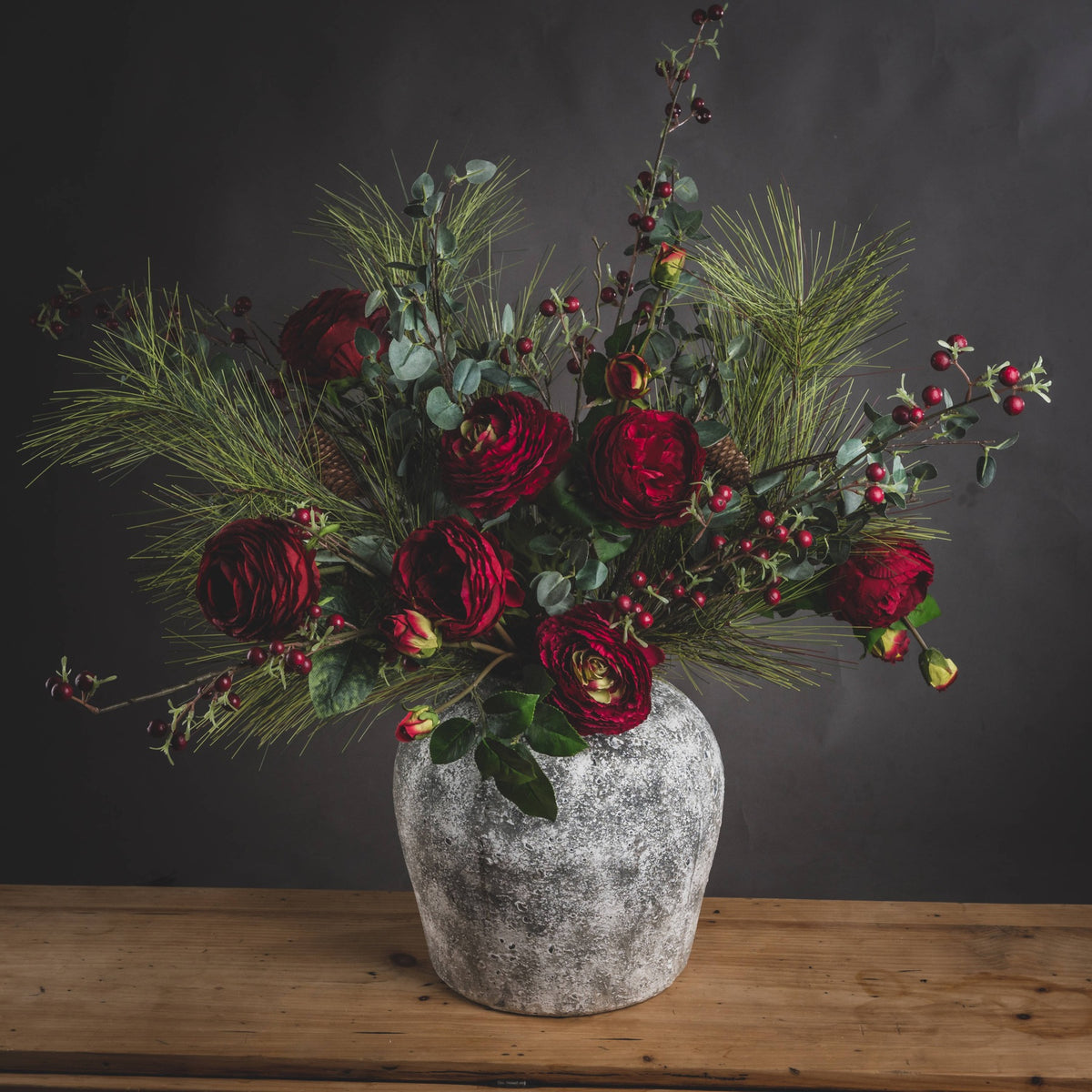 Pitted Round Vase on a wooden table with a spreading arrangement of deep red flowers, berries and green foliage against a dark wall.
