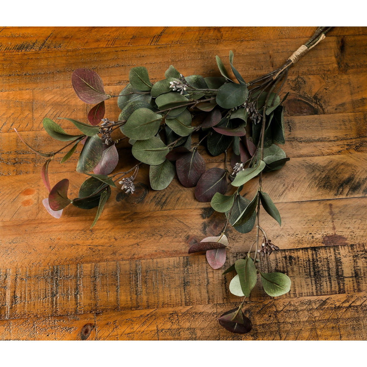 Artificial foliage bouquet lying diagonally on a wooden surface, showing veined green and burgundy leaves, pale pod-like clusters and the binding around the stems.