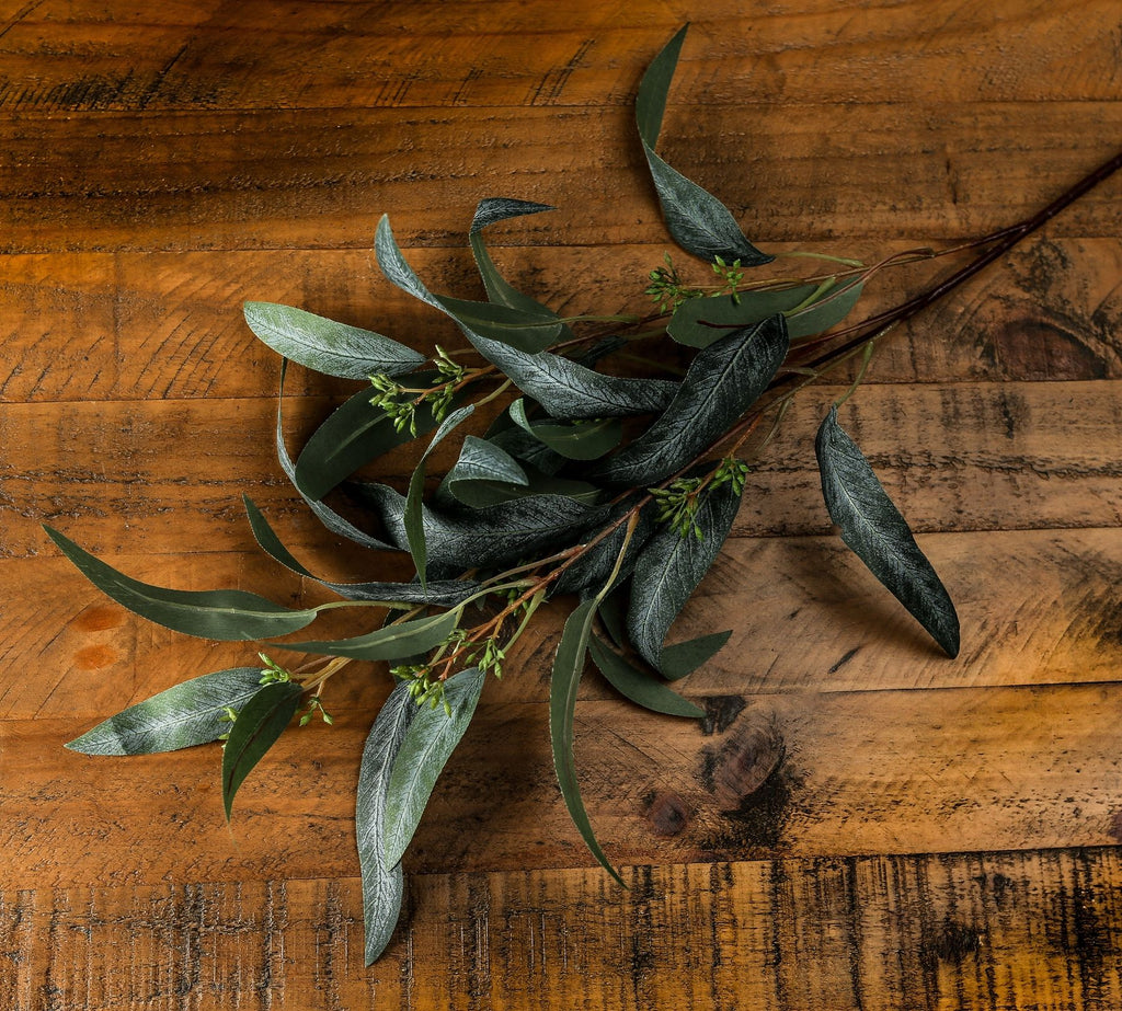 Podleaf artificial foliage laid diagonally across a wooden surface, showing curved narrow leaves and green pod-like clusters along the branches.