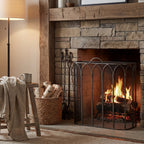 Pointed Arcade Fire Screen in front of a lit stone fireplace beneath a timber mantel, beside hearth tools and a basket of logs.