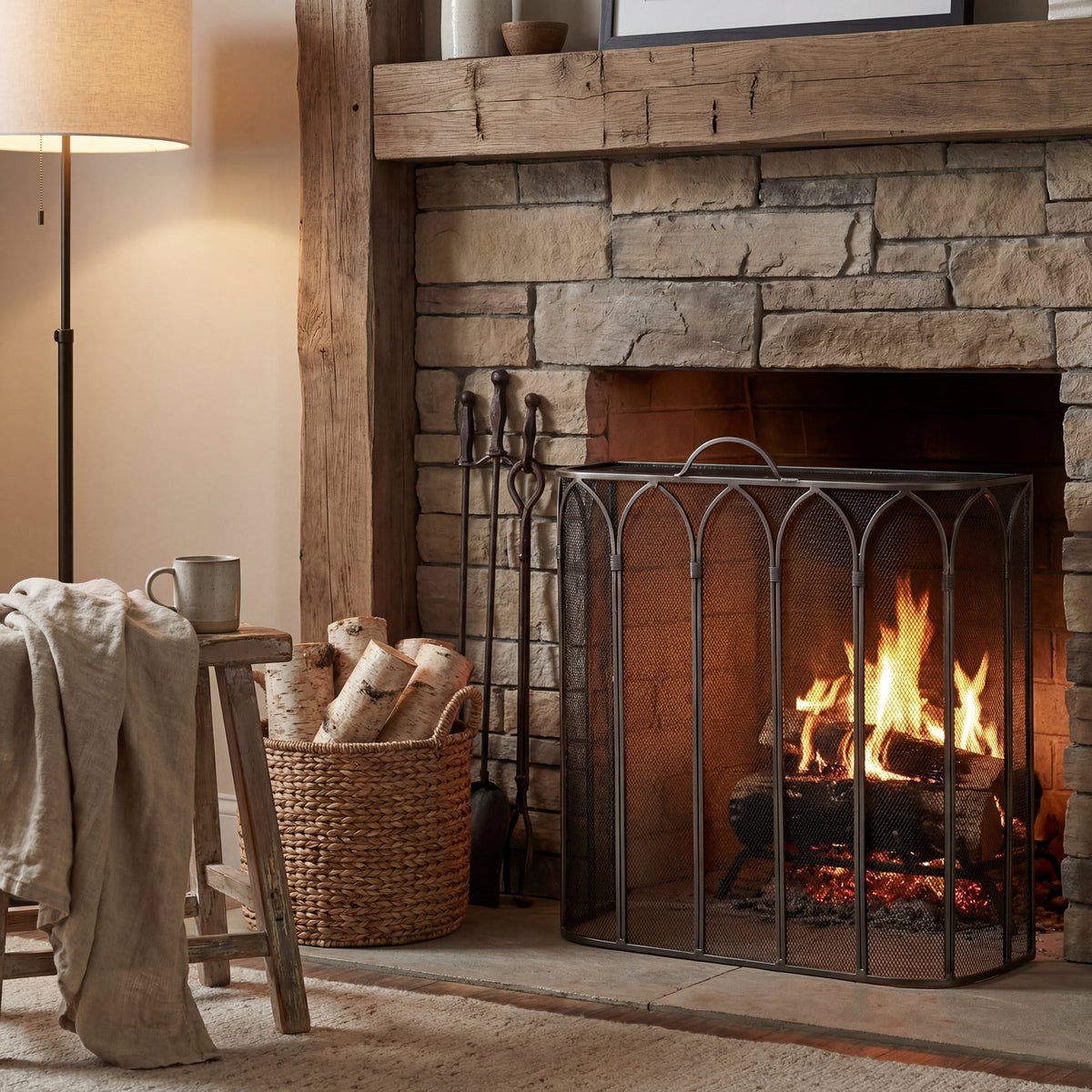 Pointed Arcade Fire Screen in front of a lit stone fireplace beneath a timber mantel, beside hearth tools and a basket of logs.