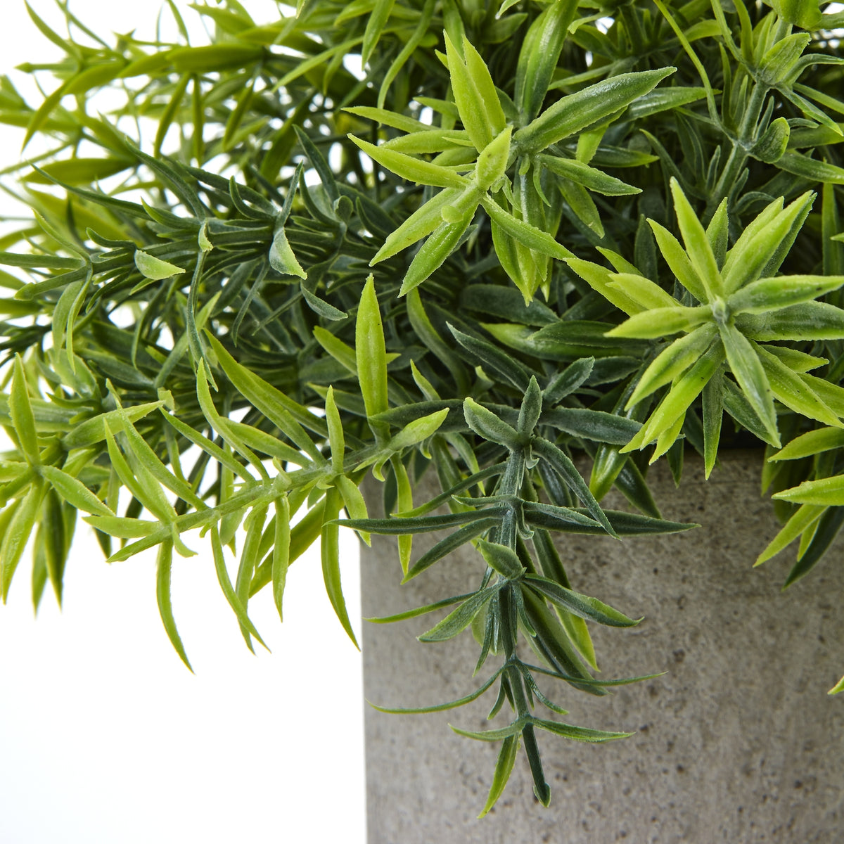 Close-up of the artificial plant's narrow, textured leaves in contrasting green tones, with a darker leafy spray hanging over the grey pot.