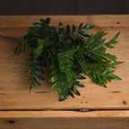 Artificial foliage bunch laid across a wooden surface, showing overlapping mid and dark green leaves with the stems extending to the left.