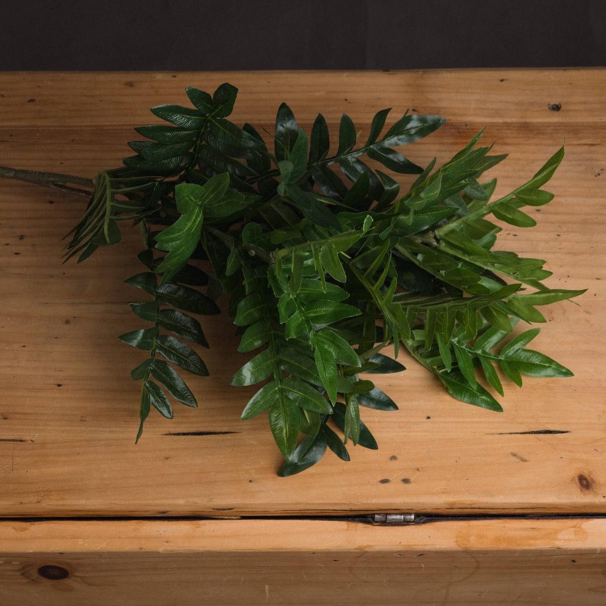 Artificial foliage bunch laid across a wooden surface, showing overlapping mid and dark green leaves with the stems extending to the left.