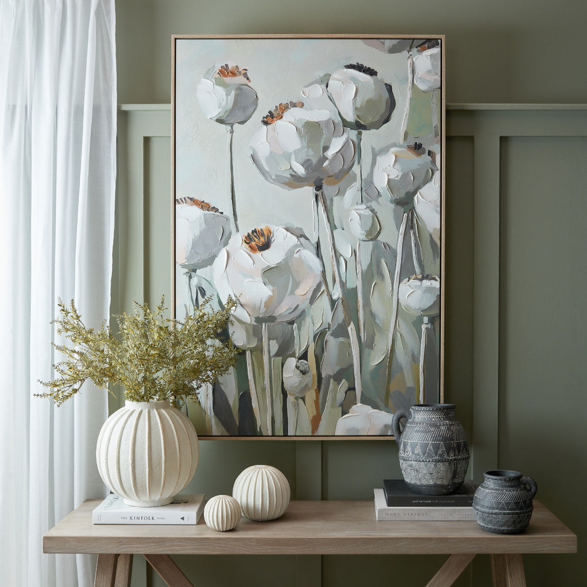 Poppy artwork displayed on a sage panelled wall above a console styled with a cream vase of foliage, rounded ornaments, dark patterned jugs and books.