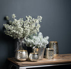Snow-effect branches arranged in a tall dark glass vase beside fuller frosted foliage and patterned metallic vessels on a wooden console against a dark wall.