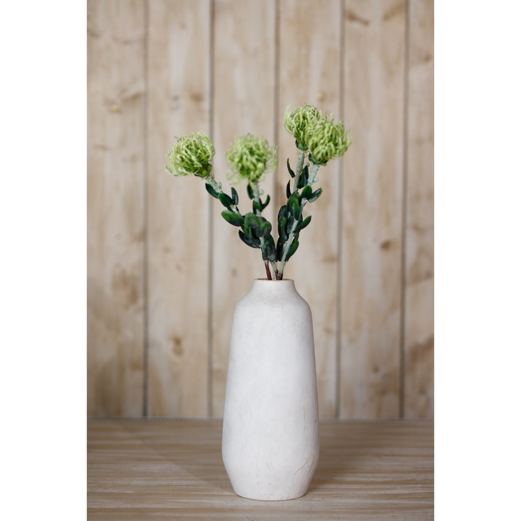 Green pincushion protea arrangement in a tall off-white vase on a wooden surface in front of vertical timber boards.