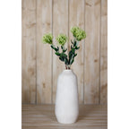 Green pincushion protea arrangement in a tall off-white vase on a wooden surface in front of vertical timber boards.