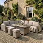 Pale cushioned woven corner seating and two stools staged on gravel beside a stone house, with coffee and pastries on the table and a throw over the right arm.