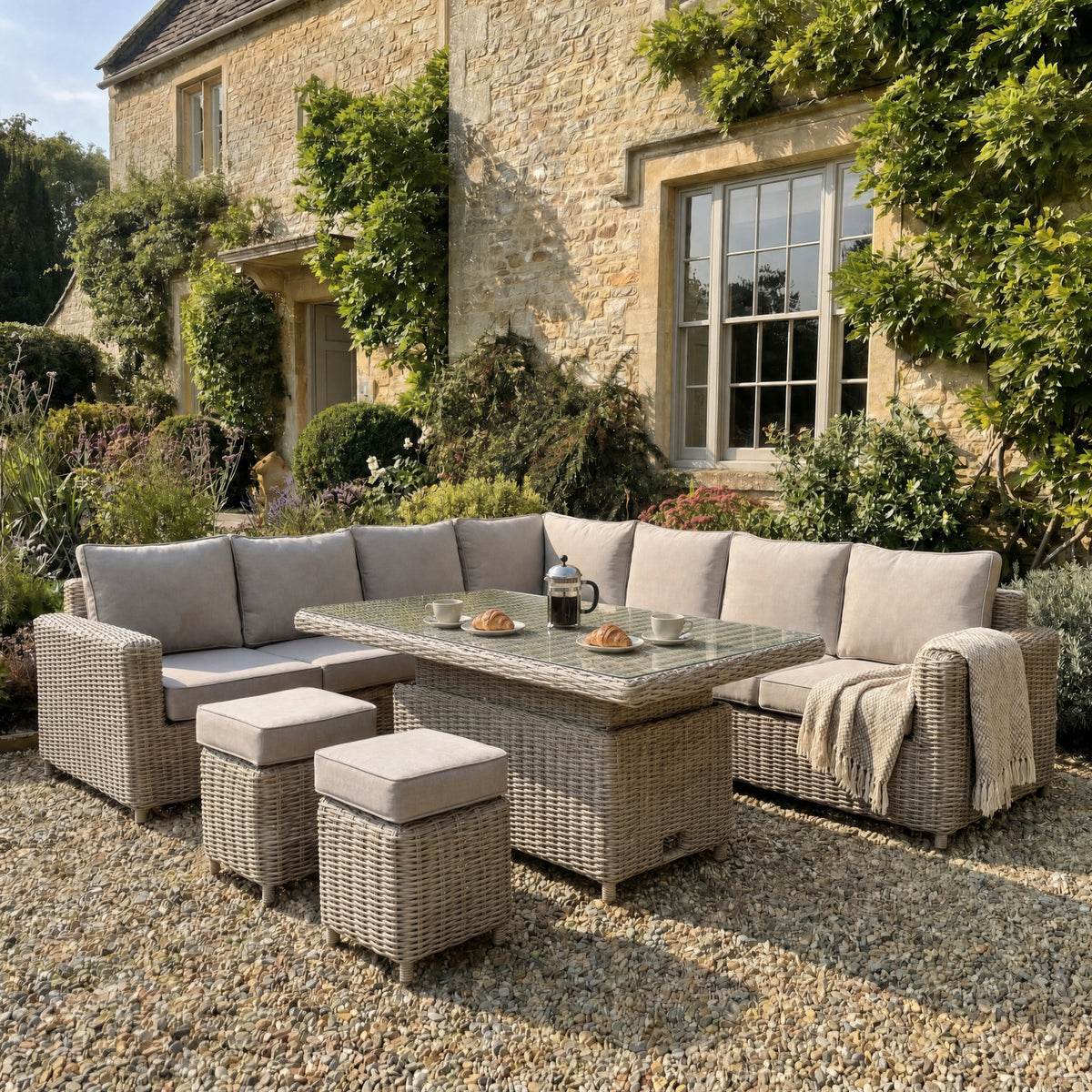 Pale cushioned woven corner seating and two stools staged on gravel beside a stone house, with coffee and pastries on the table and a throw over the right arm.