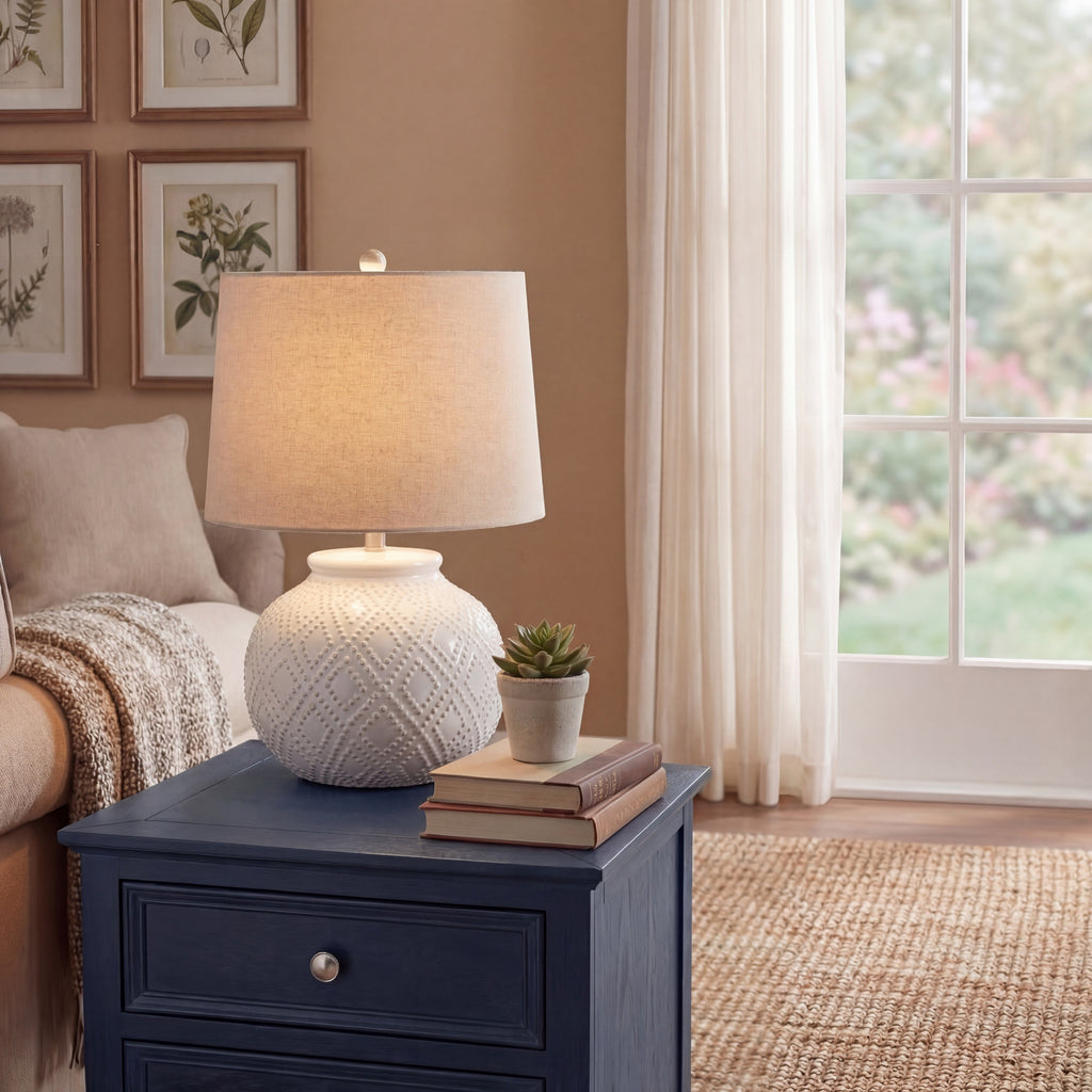 White beaded table lamp displayed on a dark blue side table beside books and a potted succulent, near a sofa and window.