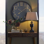 Black and gold-toned wall clock above a dark timber console, styled with a table lamp, flowers, stacked books and decorative ornaments.