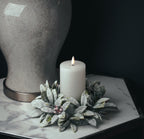 White pillar candle with an apparently illuminated flame-shaped element, styled within frosted leafy foliage and red berries beside a large crackle-finish object on a marble-effect tabletop.