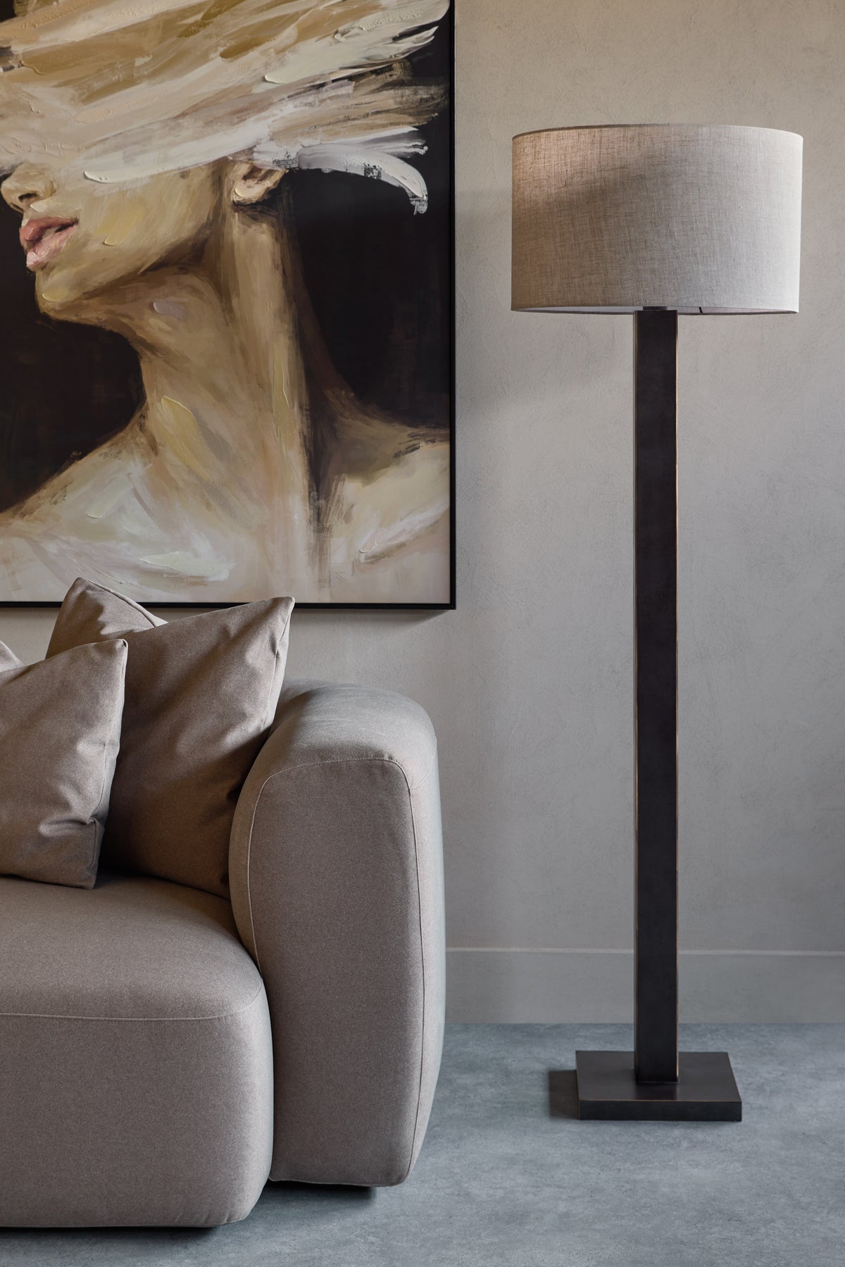 Floor lamp beside a large portrait on a pale wall, with a taupe sofa and neutral cushions beneath the portrait.