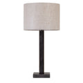 Front view of a dark bronze table lamp with a tall rectangular stem, square base and natural-toned drum shade on white.
