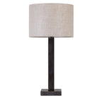 Front view of a dark bronze table lamp with a tall rectangular stem, square base and natural-toned drum shade on white.