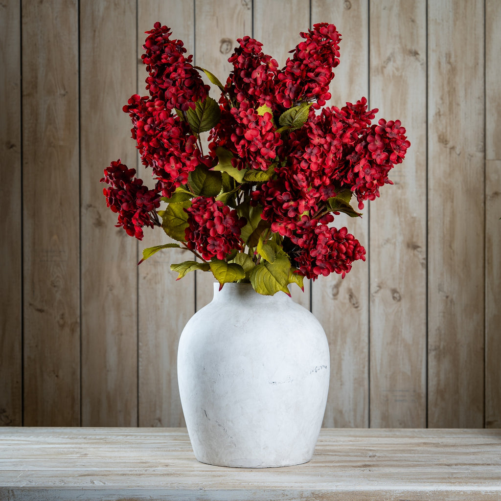 Red artificial flowers and green foliage arranged in a pale rounded vase on a wooden surface against vertical wood panelling.