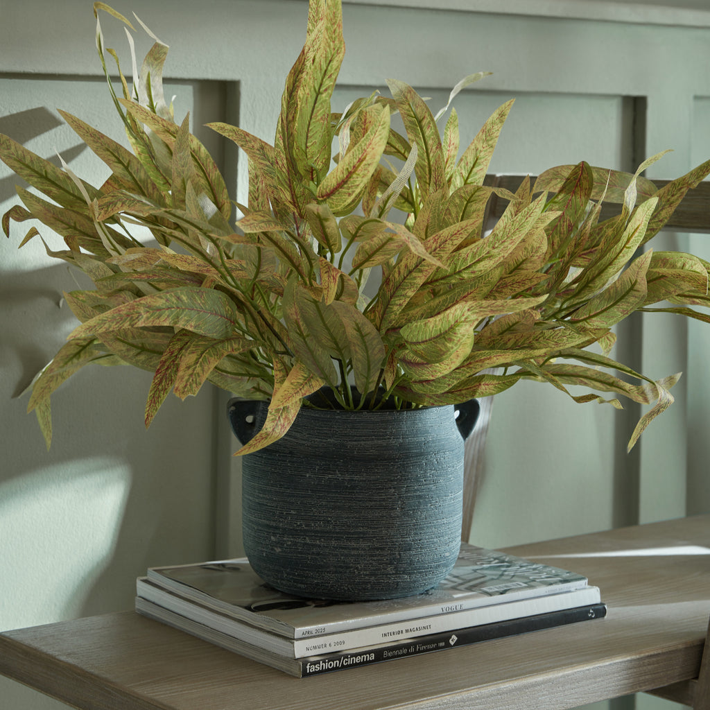 Styled foliage arrangement in a dark textured handled vase on stacked magazines and a wooden surface, against a pale green panelled wall.