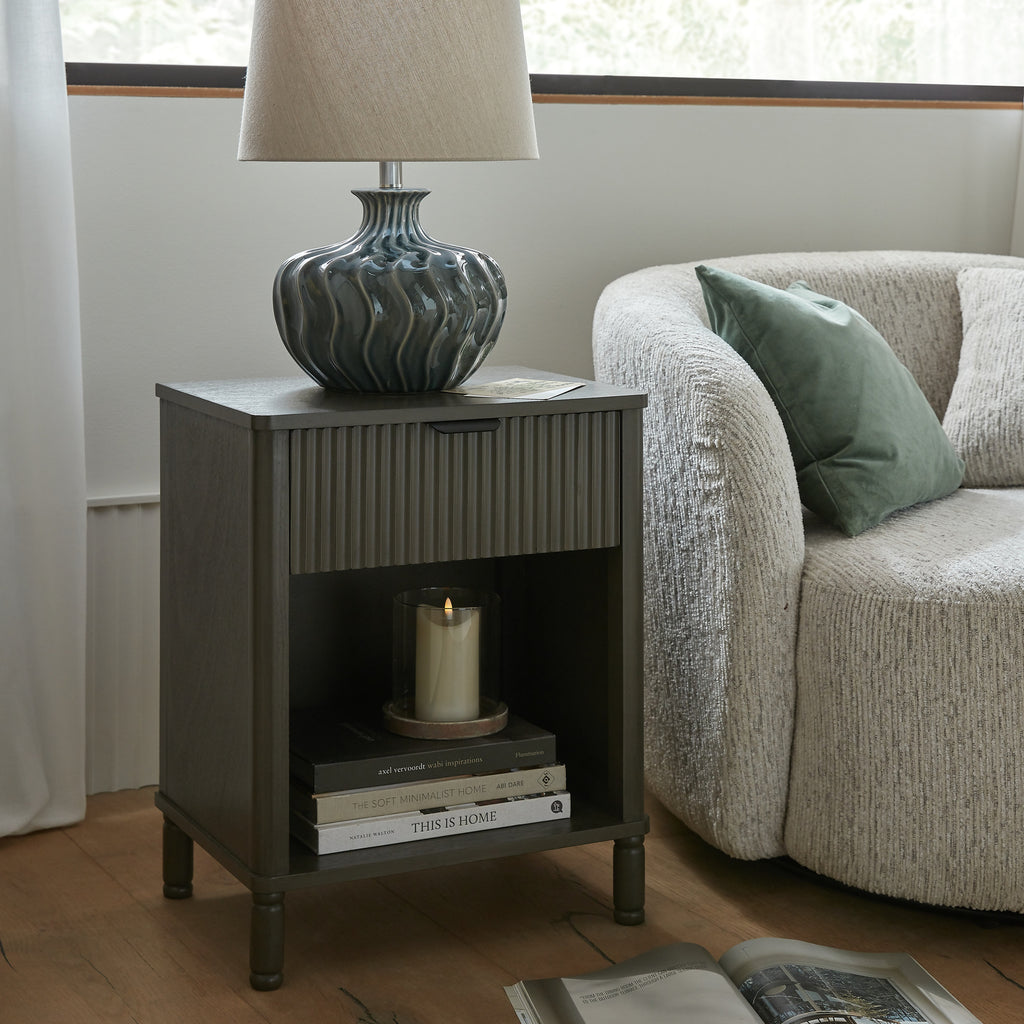 Cabinet beside cream upholstered seating, with a ceramic table lamp on top and stacked books and a glass candle holder in the cubby.