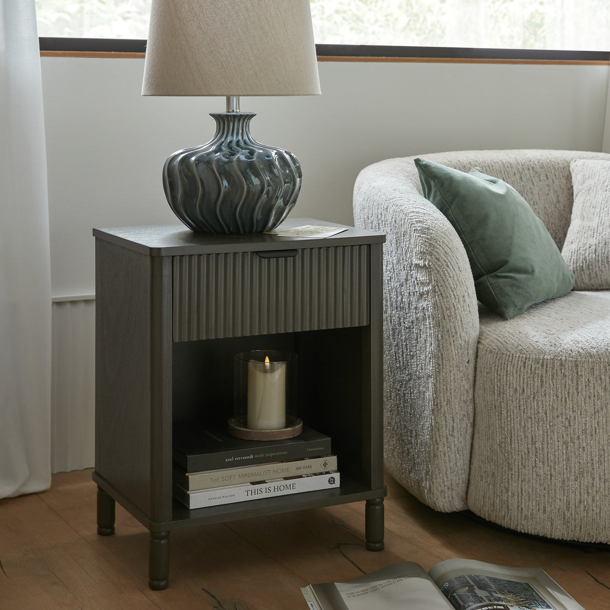 Cabinet beside cream upholstered seating, with a ceramic table lamp on top and stacked books and a glass candle holder in the cubby.