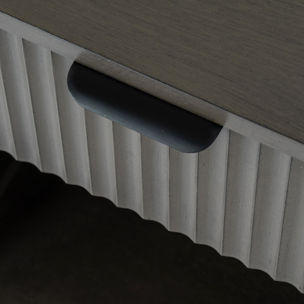 Close-up of the dark cup-shaped drawer pull beneath the wooden top, with vertical reeding across the drawer front.