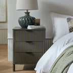Reeded bedside table beside a bed with white textured bedding, styled with a green lamp, stacked books and an amber glass vessel.