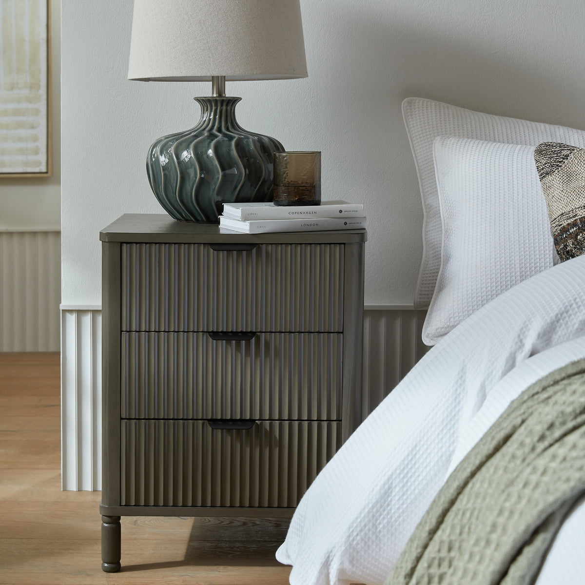 Reeded bedside table beside a bed with white textured bedding, styled with a green lamp, stacked books and an amber glass vessel.