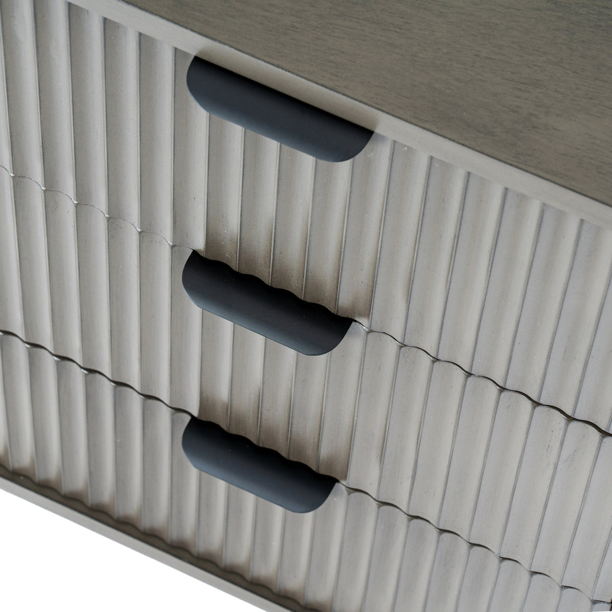 Close-up looking down across the bedside table's vertically ridged drawer fronts and three dark cup-shaped pulls beneath the plain top.