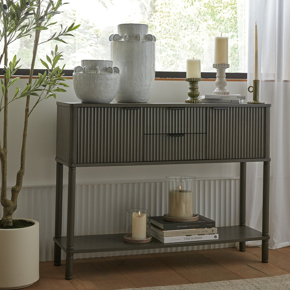 Reeded console table beneath a window, styled with pale decorative vessels and candles on top and books and glass candle holders on the lower shelf, beside a potted tree.