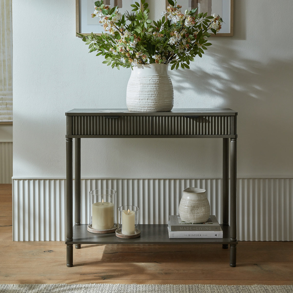 Console table against a pale panelled wall, styled with a white vase of flowering branches on top and glass candle holders, books and a smaller white vase on the lower shelf.