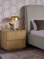 Oak nightstand beside a grey upholstered bed, styled with a gold-toned lamp, book, small vase and dish against patterned wallpaper.