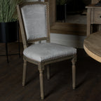 Pale grey stud-edged dining chair beside a round brown table, with a large framed mirror behind it and a tall black planter to the left.