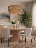 Round ribbed pedestal dining table surrounded by pale upholstered chairs, styled with flowers and candles beneath a large woven pendant.