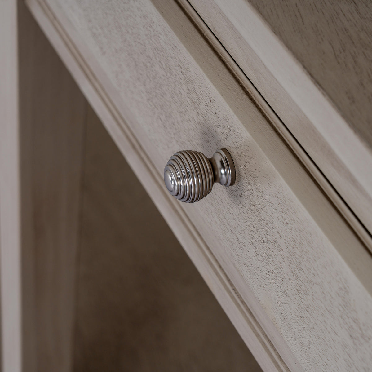 Close-up of a rounded silver-coloured ribbed knob against the pale grained drawer front and its bevelled border.