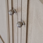 Close-up of the cabinet's two silver-coloured ribbed knobs beside raised door mouldings and the pale grey-beige grained finish.