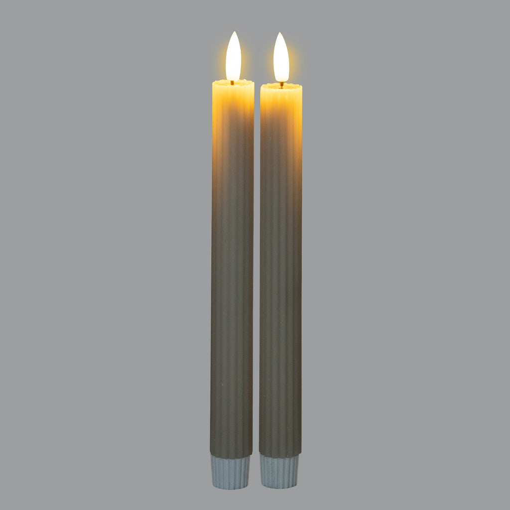 Two ribbed candles photographed with bright flame-shaped tips and a golden glow around their tops against a grey background.