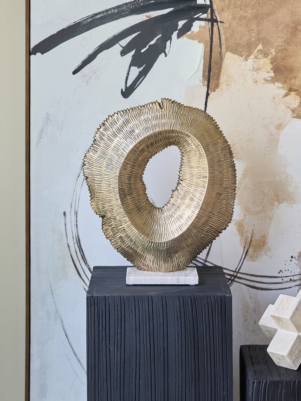 Antique gold hollow sculpture displayed on a black ridged plinth in front of black, white and gold abstract artwork.