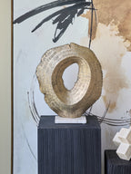 Antique gold hollow sculpture displayed on a black ridged plinth in front of black, white and gold abstract artwork.