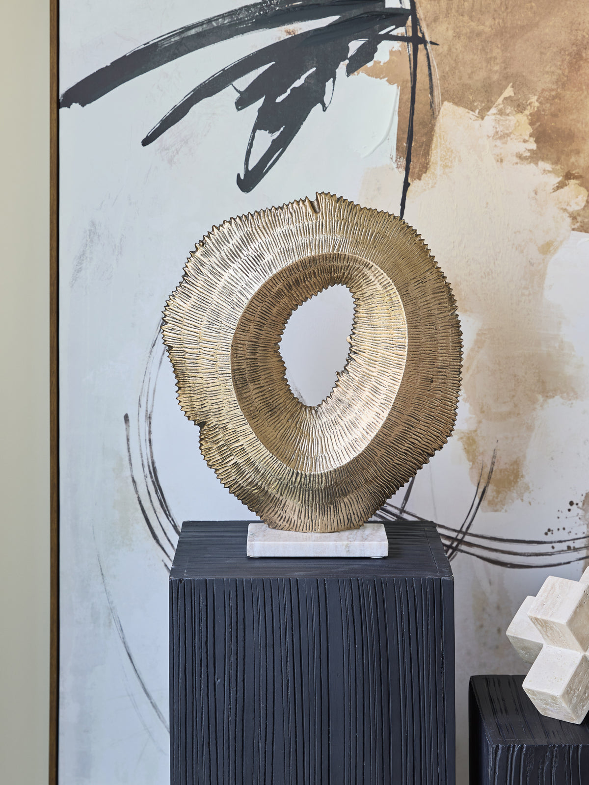 Antique gold hollow sculpture displayed on a black ridged plinth in front of black, white and gold abstract artwork.