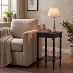Illuminated table lamp on a dark wooden side table beside a pale upholstered armchair, with books, a mug and surrounding plants.