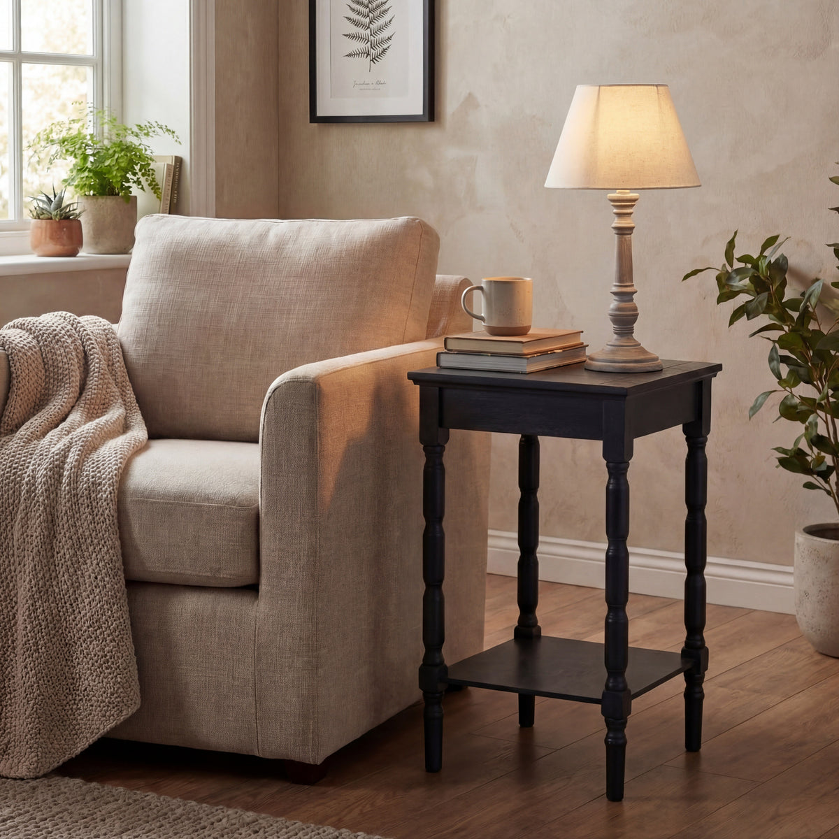 Illuminated table lamp on a dark wooden side table beside a pale upholstered armchair, with books, a mug and surrounding plants.