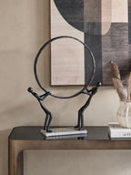 Ring-bearing figures sculpture on a dark console beneath geometric artwork, beside books and a pale vase of dried stems.