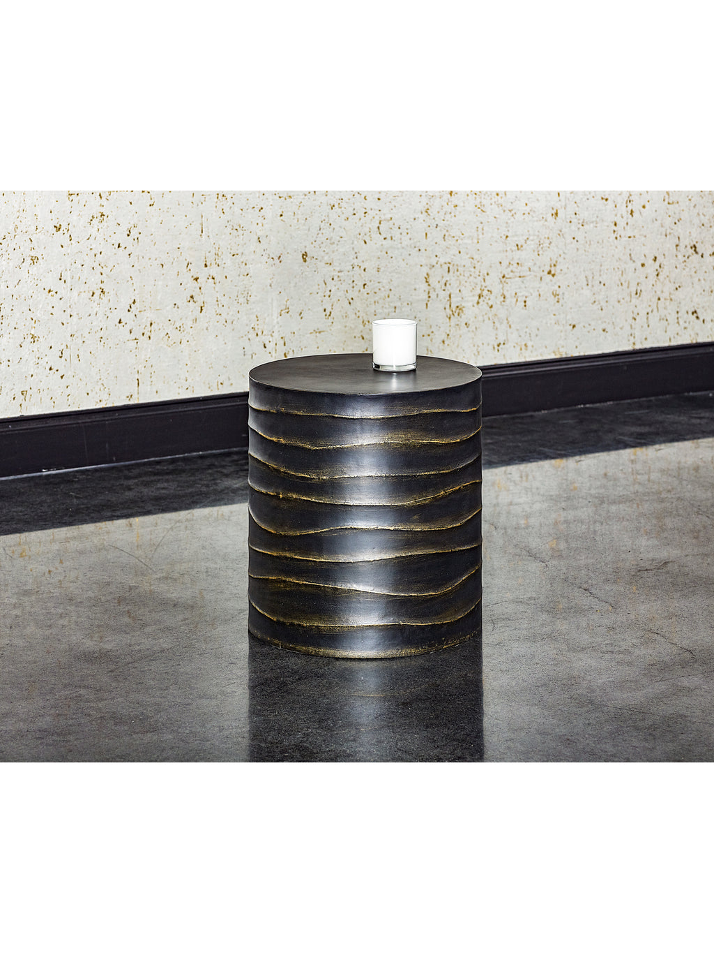 Rippled black drum side table holding a white candle in a glass vessel, beside a pale textured wall on a reflective dark floor.