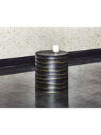 Rippled black drum side table holding a white candle in a glass vessel, beside a pale textured wall on a reflective dark floor.