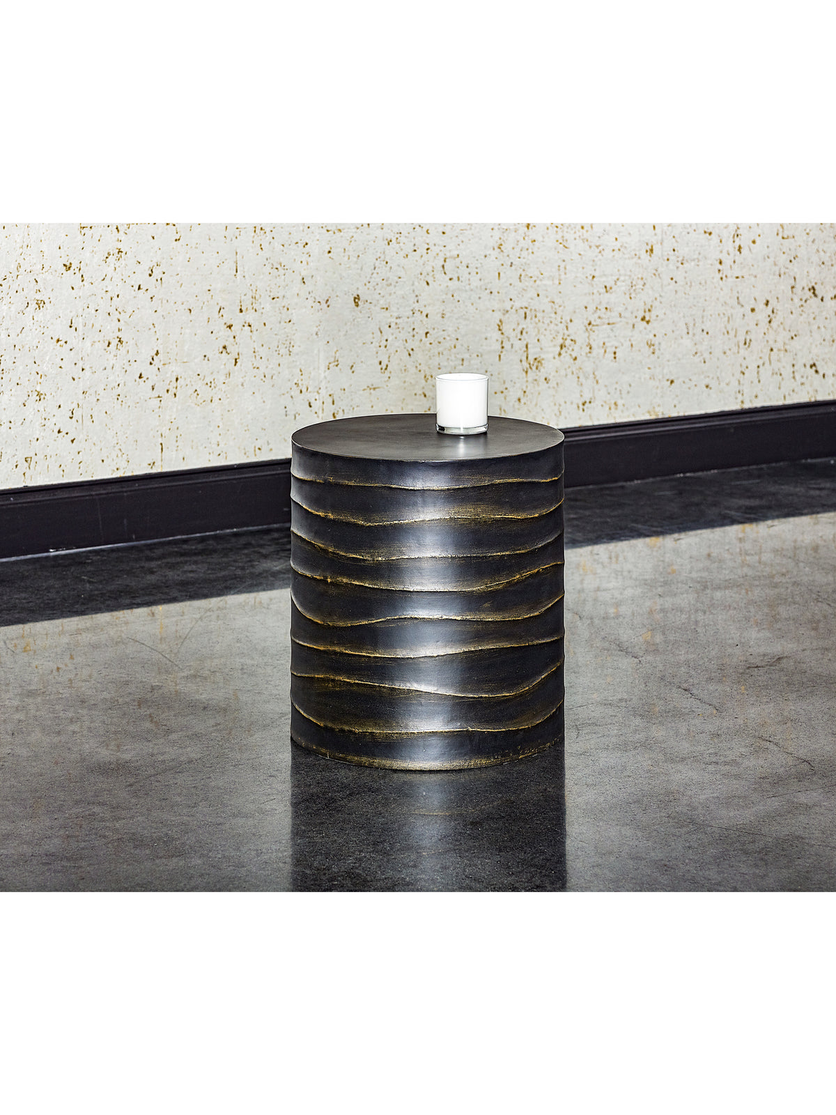 Rippled black drum side table holding a white candle in a glass vessel, beside a pale textured wall on a reflective dark floor.